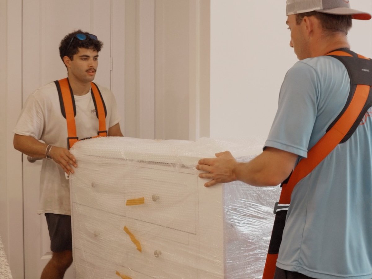 Two McCuller Moving crew members using lifting straps to carry a wrapped dresser through an apartment
